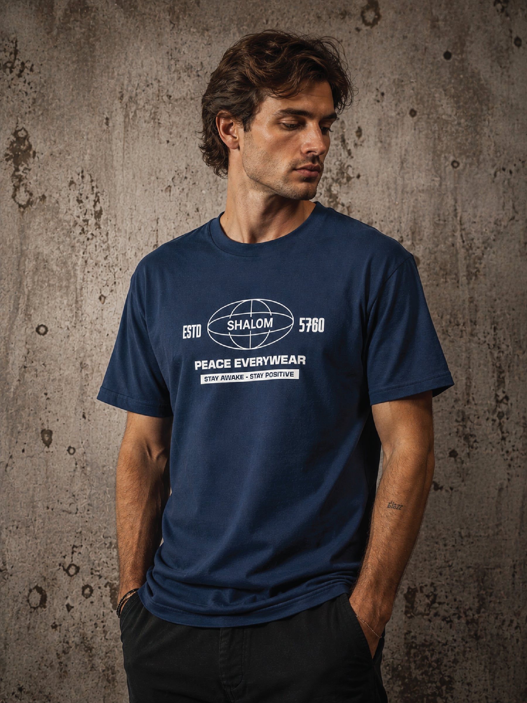 A man wearing a navy blue graphic t-shirt with a white 'Shalom Peace Everywear' globe logo on the chest, standing against a textured concrete wall.
