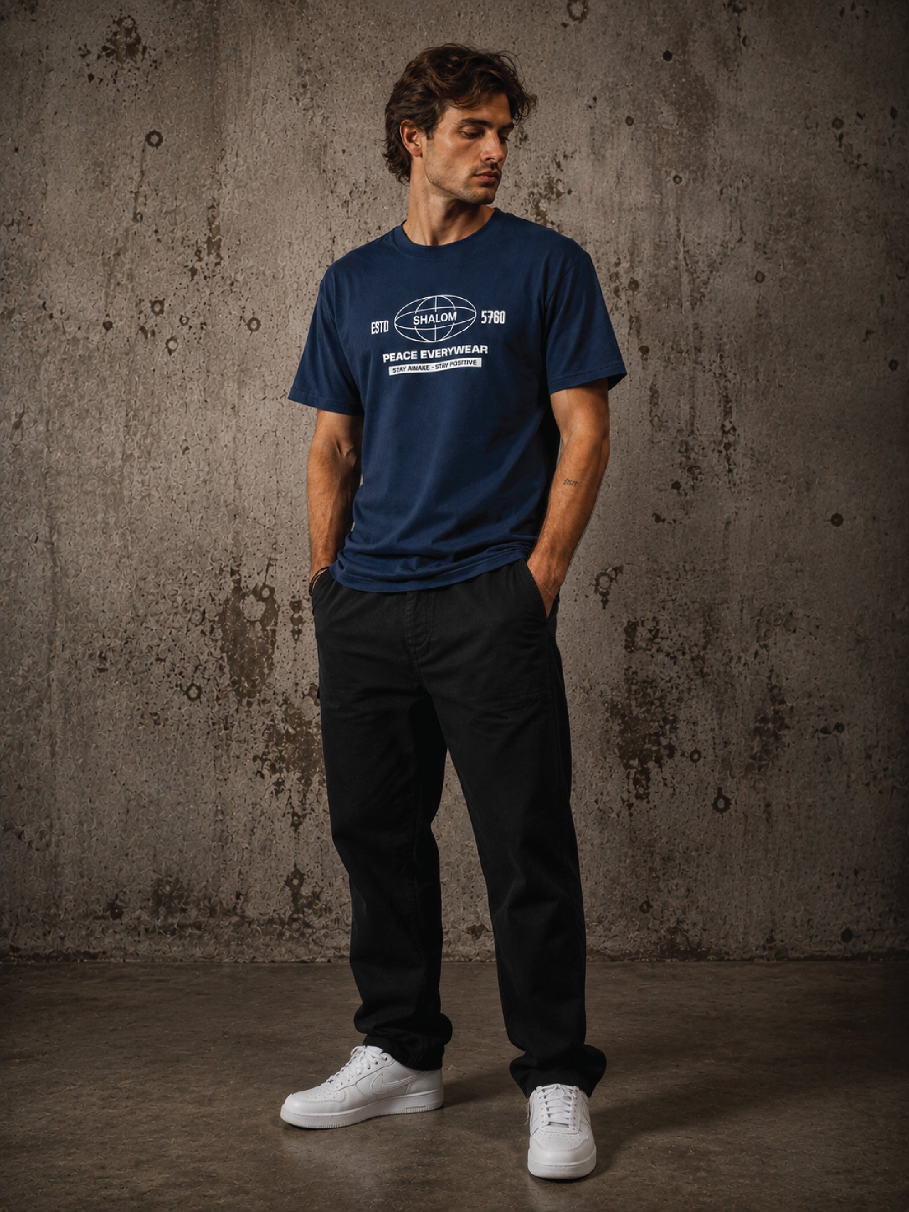 A man wearing a navy blue graphic t-shirt, black pants, and white sneakers stands against a textured concrete wall.
