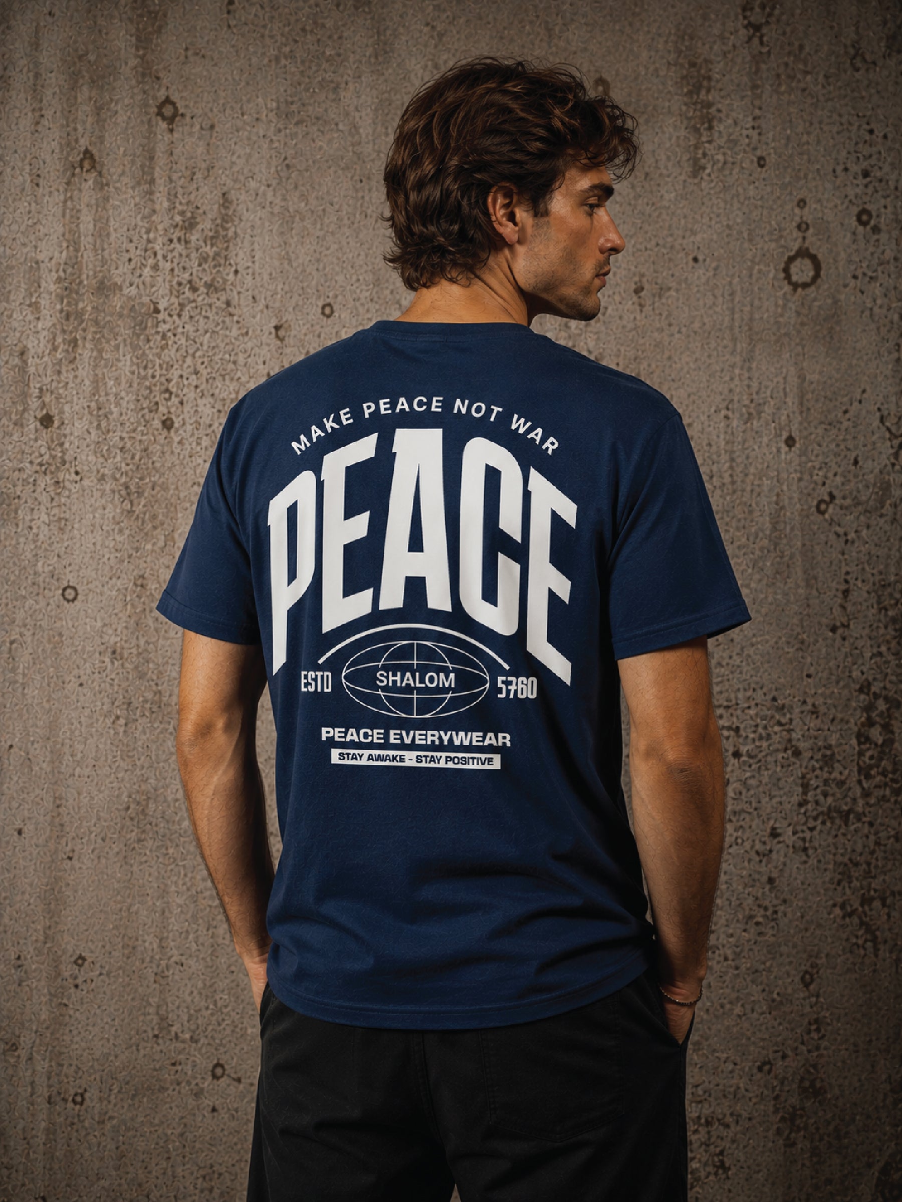A man viewed from the back wearing a navy blue t-shirt with a large white graphic print on the back that reads 'MAKE PEACE NOT WAR PEACE ESTD 1970 SHALOM 1979 PEACE EVERYWEAR'. He is standing against a textured concrete wall.