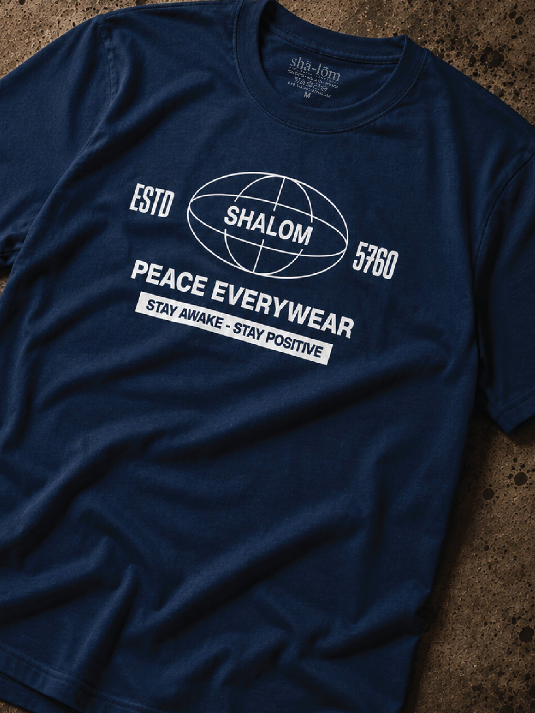 A navy blue crewneck t-shirt with a white graphic print on the front. The graphic features a globe design with the word 'SHALOM' inside, flanked by 'ESTD 5760' and 'PEACE EVERYWEAR' above a banner reading 'STAY AWAKE - STAY POSITIVE'.