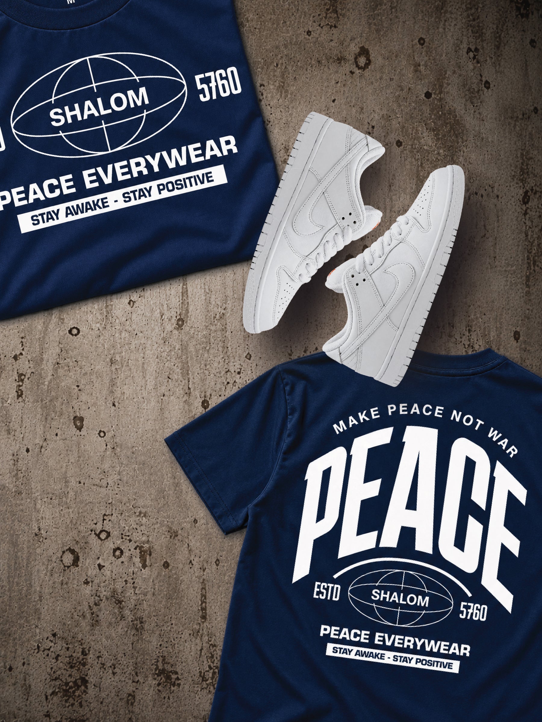 A styled flat-lay of navy blue t-shirts and white sneakers on a textured concrete surface. One t-shirt shows the front with a white 'SHALOM PEACE EVERYWEAR' graphic, another shows the back with a large 'PEACE' graphic, and a pair of white low-top sneakers is placed between them.